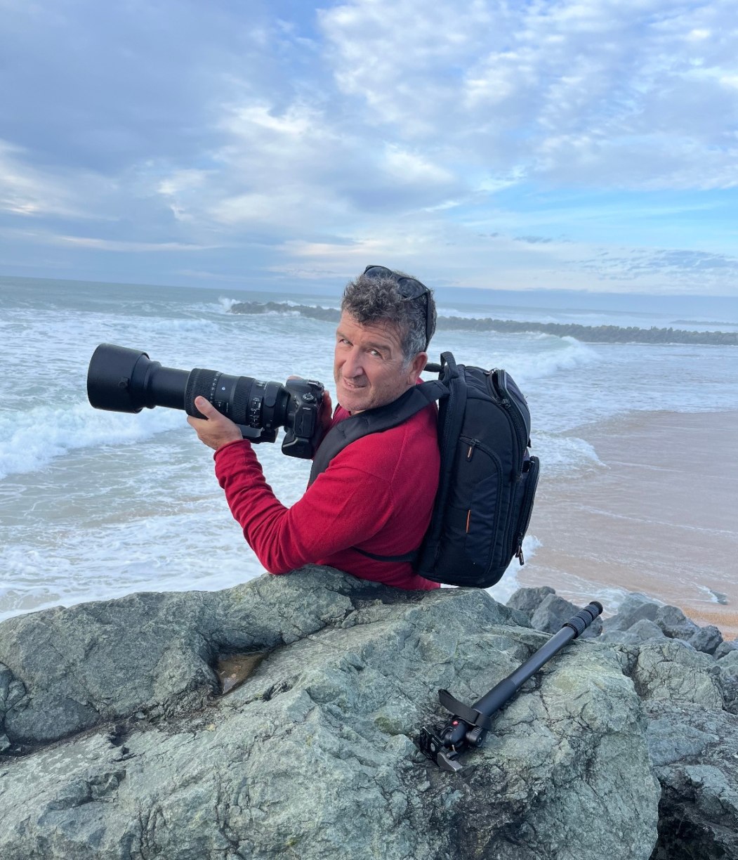 Éric Zudaire, appareil photo et téléobjectif en main, assis sur les rochers face à l'océan